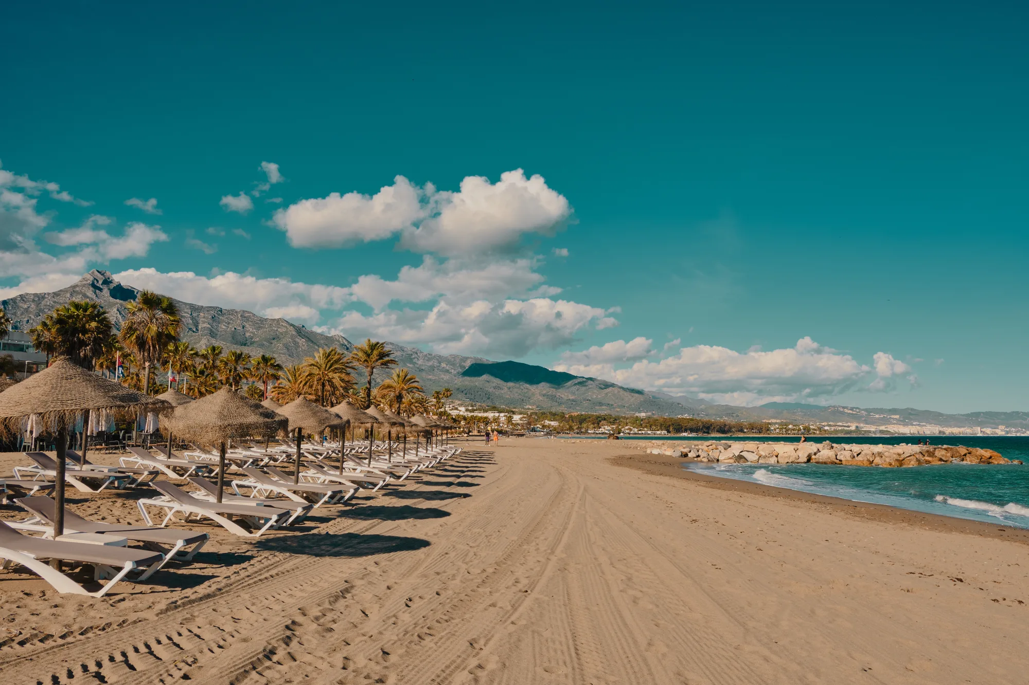 Peščana plaža sa ležaljkama i suncobranima od trske, lukobran i planine u pozadini po sunčanom danu
