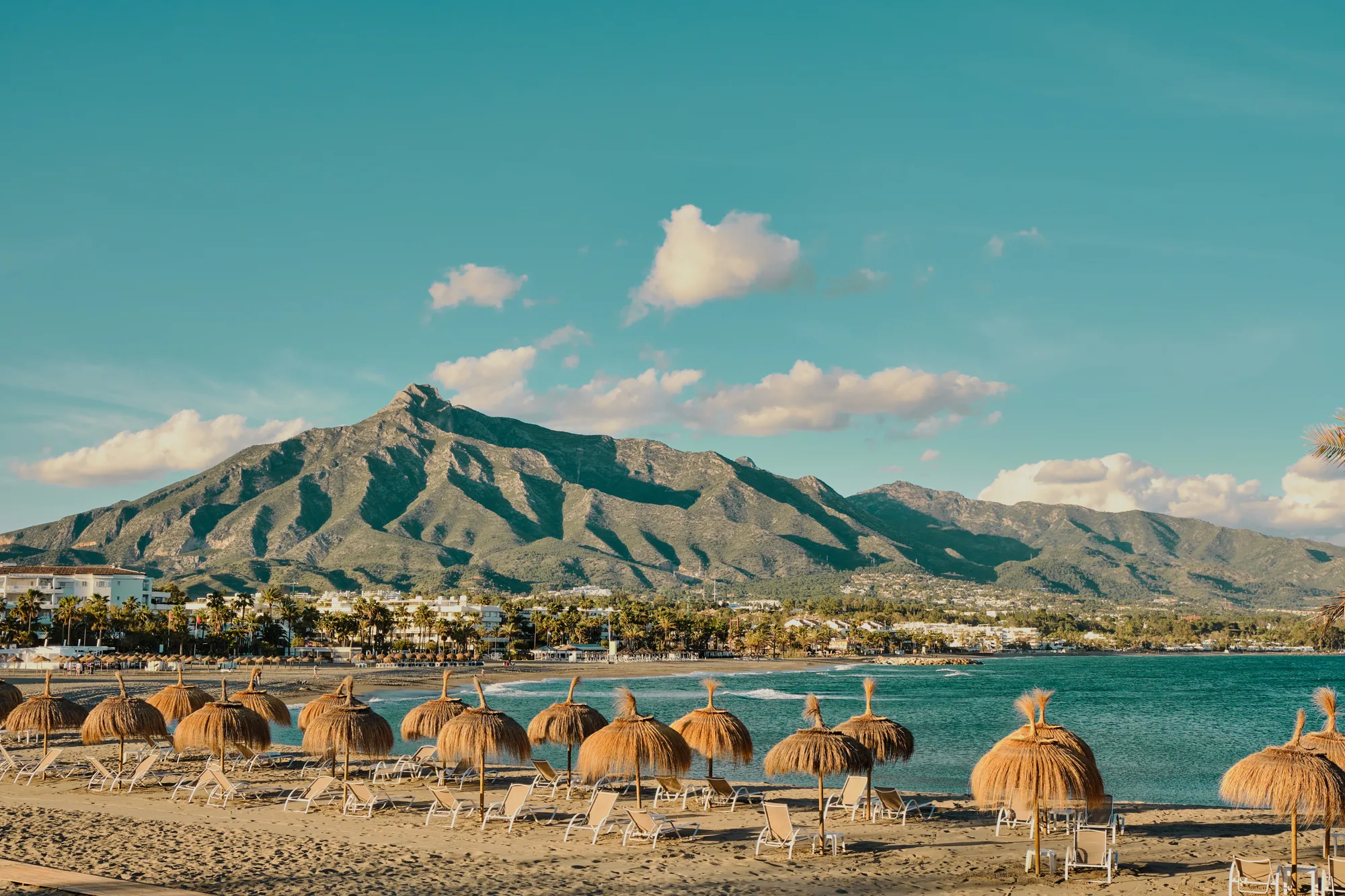 Plaža sa suncobranima od trske, tirkizno more i planina La Concha (Sierra Blanca) iznad Marbelle