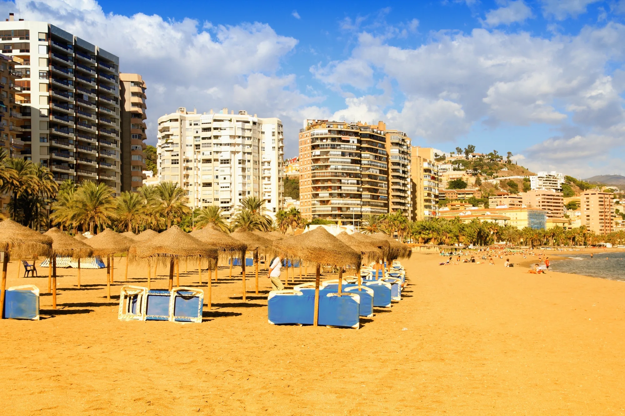 Peščana gradska plaža sa plavim ležaljkama i suncobranima od trske, palme na šetalištu i visoke stambene zgrade u toplom popodnevnom svetlu