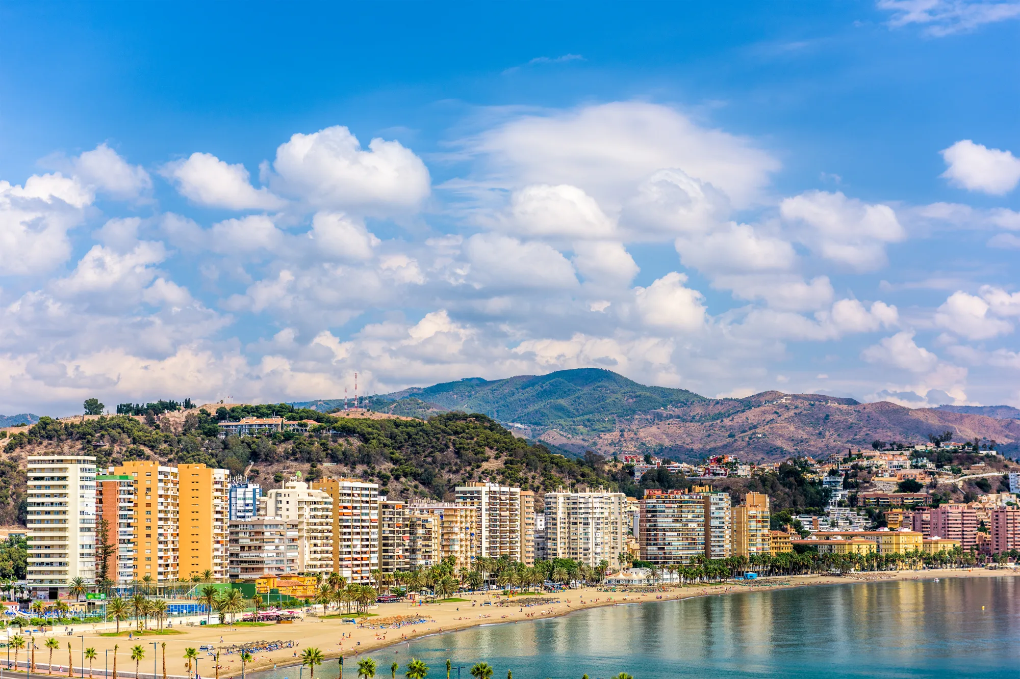 Gradska plaža sa palmama uz šetalište, niz visokih zgrada, brdo Gibralfaro i planine pod oblacima