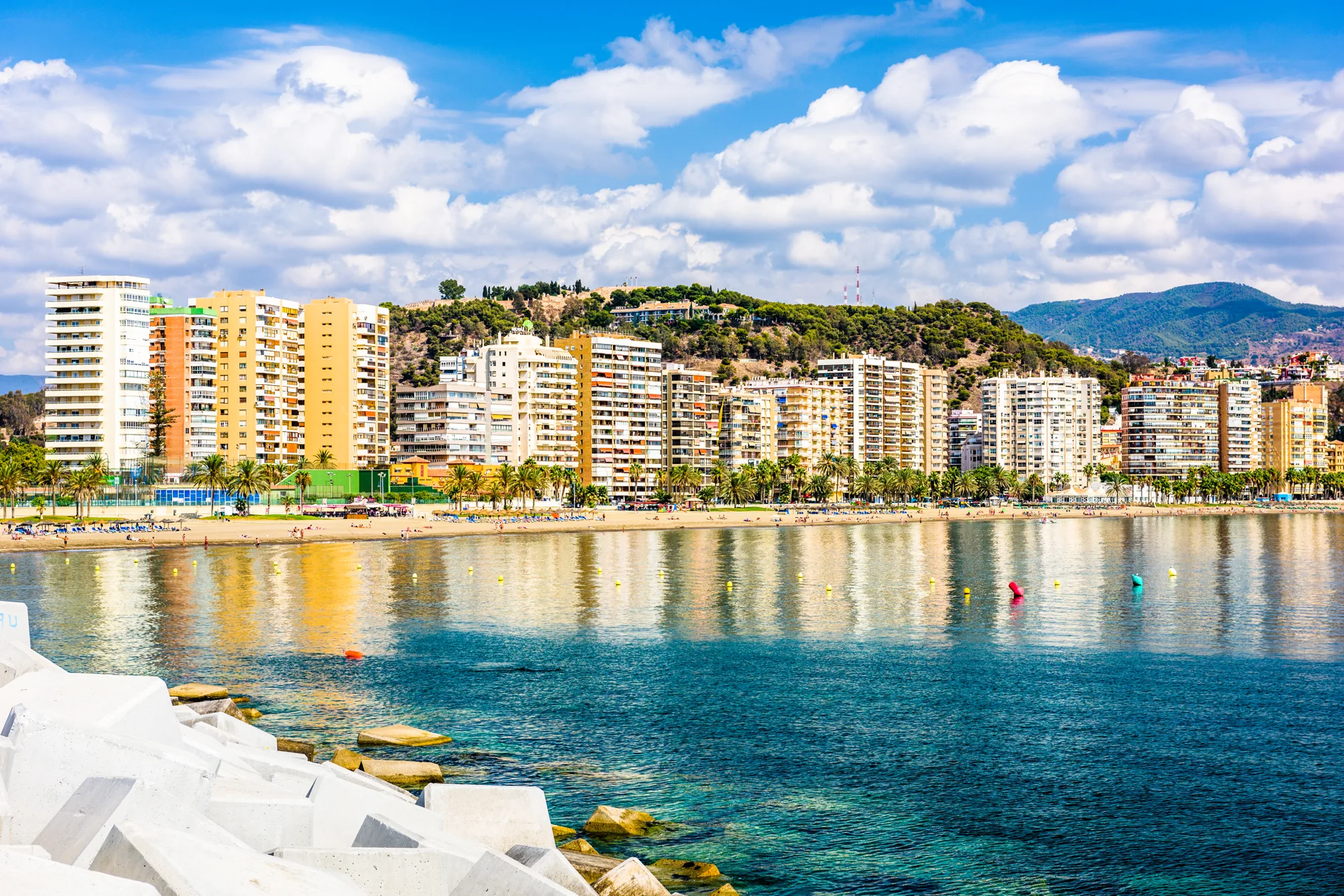 Tirkizno more i gradska plaža sa nizom visokih zgrada uz šetalište i palmama, brdo Gibralfaro i planine iza grada