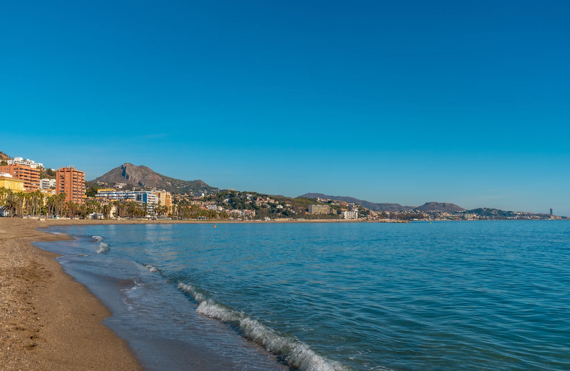 Peščana plaža sa talasima koji zapljuskuju obalu, zgrade i palme uz šetalište i brda iza grada pod vedrim nebom