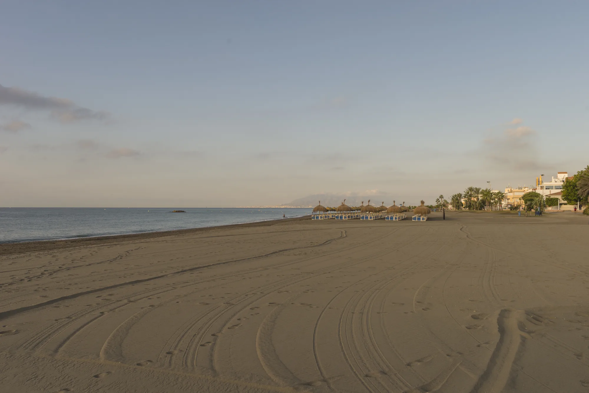 Široka plaža sa tamnim peskom i tragovima na pesku, mirno more i par suncobrana u daljini, bez gužve