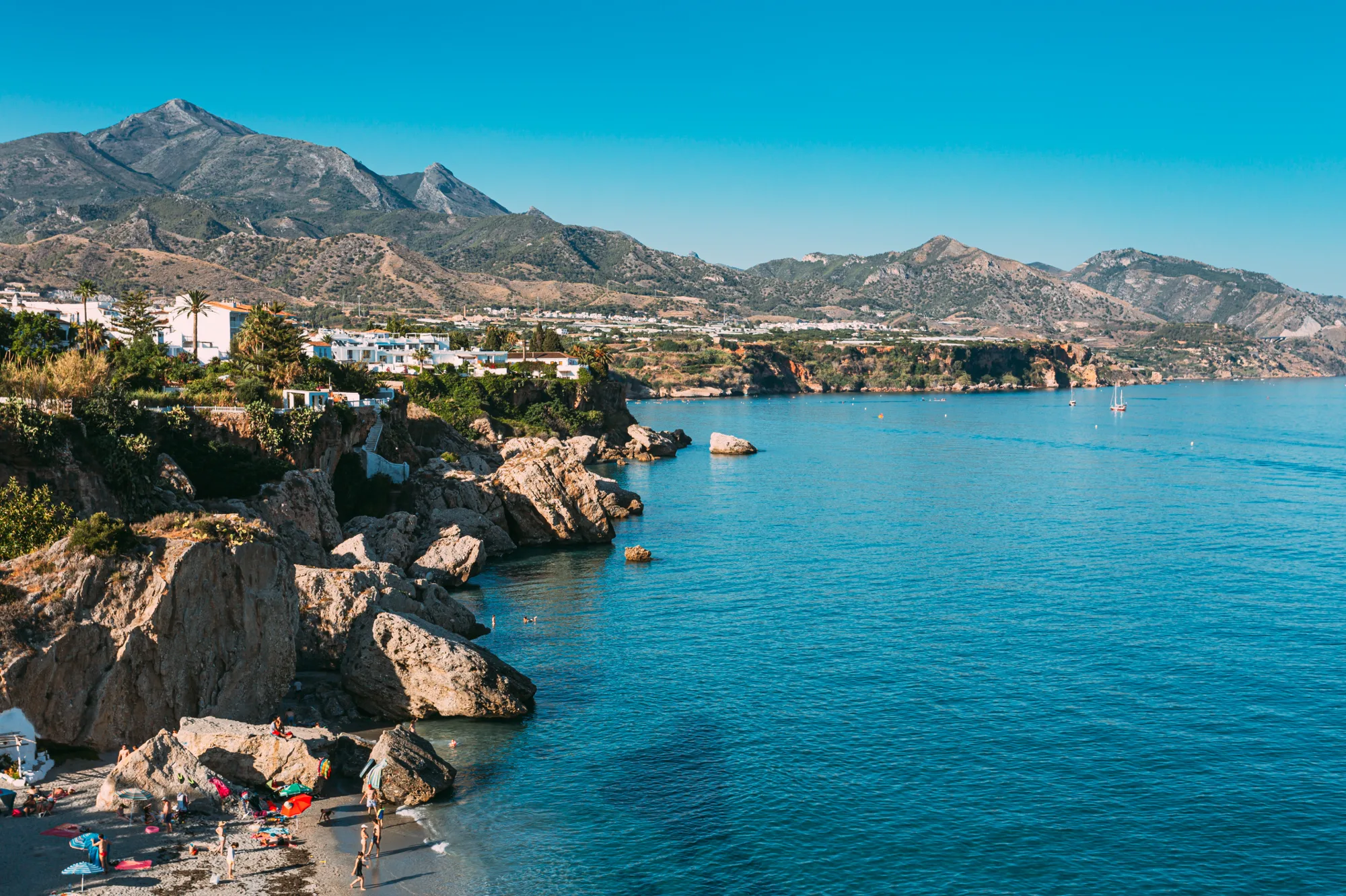 Široki pogled na stenovitu obalu sa liticama, tirkiznim morem, malom plažom ispod stena i planinama iznad grada