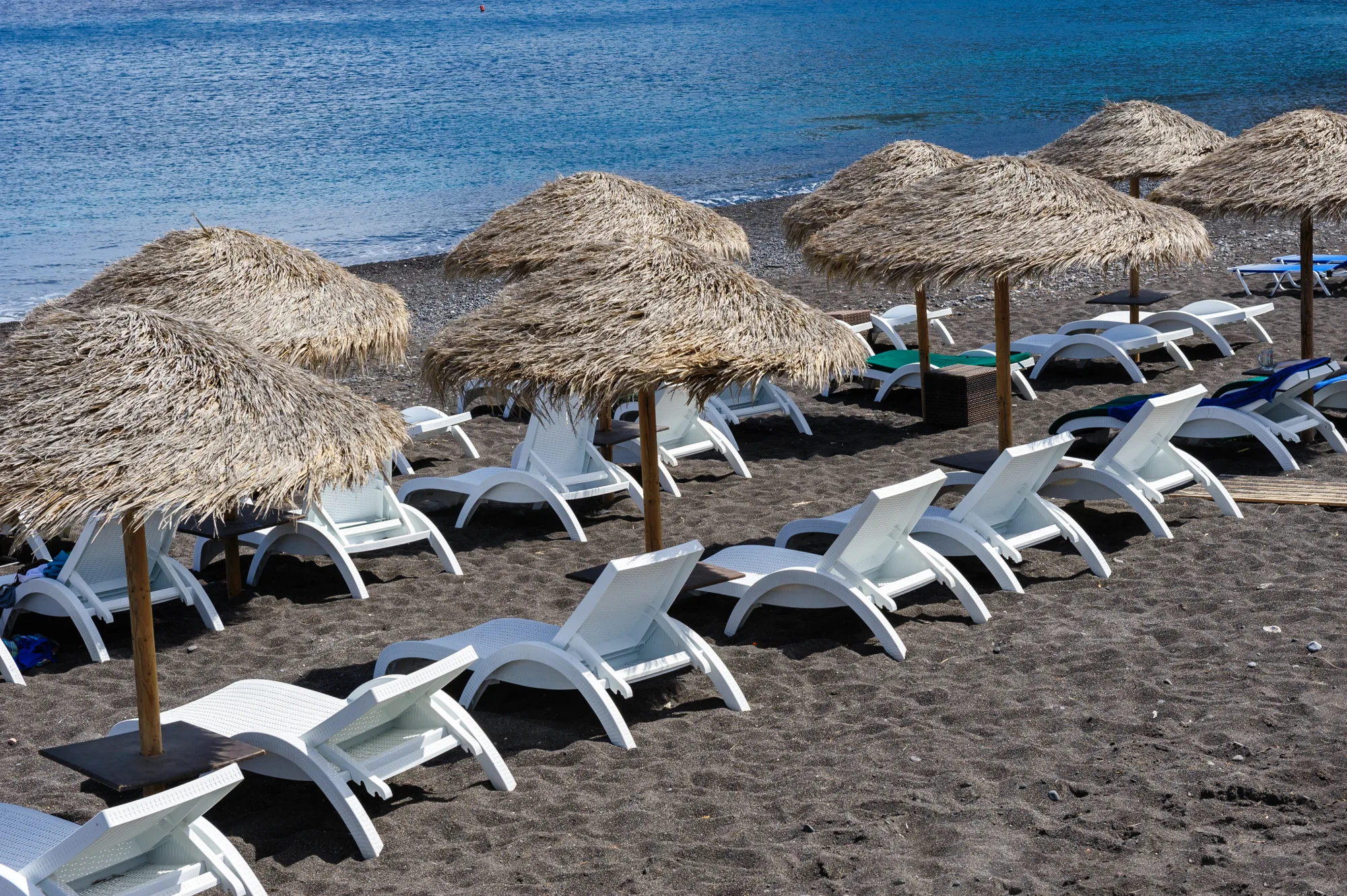 Redovi belih ležaljki pod suncobranima od trske na plaži tamnog peska uz plavo more